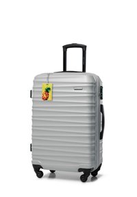 Silver hard-shell suitcase with four wheels, black retractable handle, side handles, and a yellow pineapple luggage tag attached.