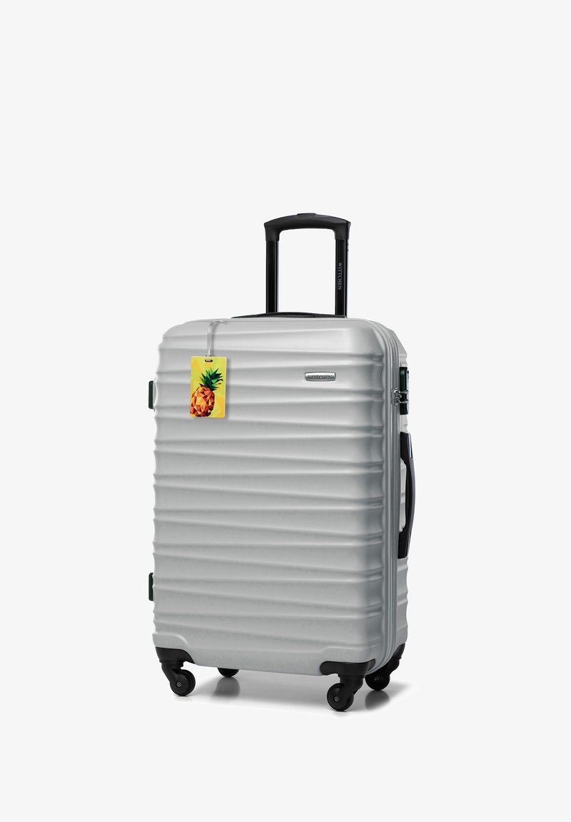 Srebrna twarda walizka na czterech kółkach, czarny wysuwany uchwyt, boczne uchwyty i załączona żółta zawieszka w kształcie ananasa.