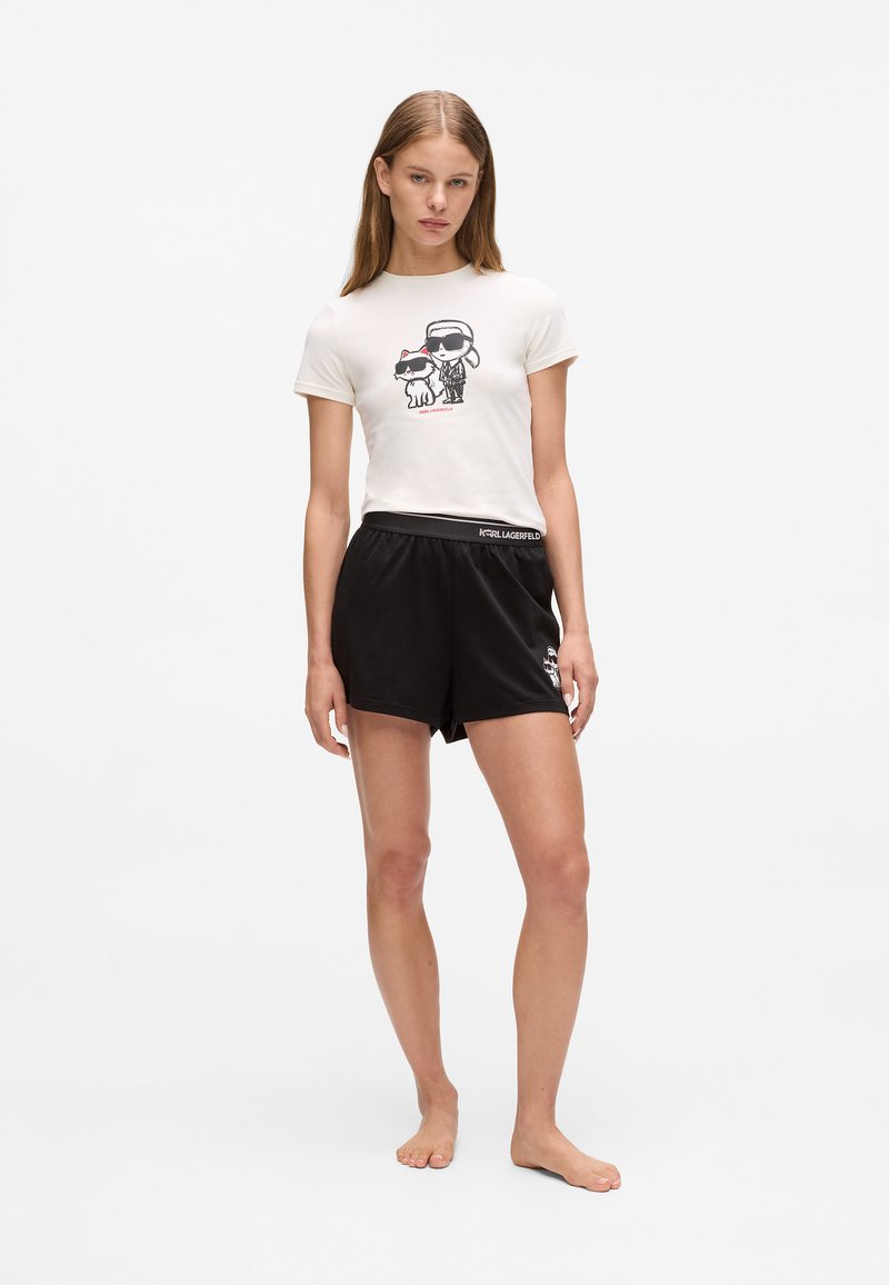 Vit grafisk t-shirt med en tecknad design, kombinerad med svarta shorts. Shortsen har en elastisk midja med en logotyp.