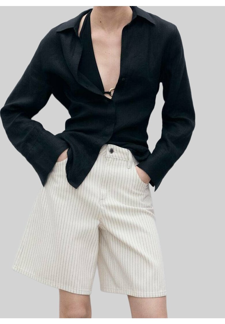 Personne portant une chemise noire à manches longues sur un top noir et un short blanc rayé jusqu'aux genoux, les mains dans les poches.