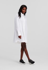 Camicia bianca a maniche lunghe con bottoni, orlo arrotondato e vestibilità oversize, abbinata a scarpe da ginnastica nere con suole bianche e lacci decorativi.