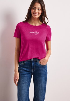 Femme souriante, portant un t-shirt à manches courtes rose vif avec le texte « Atelier Soleil », associé à un jean bleu, main dans la poche.