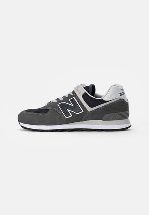 Mørkegrå og sort New Balance-sneaker med hvid sål og logo, snøredesign og polstret hæl på en ensartet hvid baggrund.