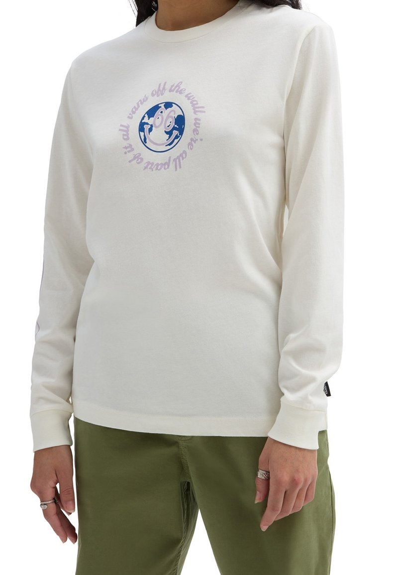 Person in einem cremefarbenen Langarmshirt mit einem blauen lächelnden Globus und lila Text, kombiniert mit olivgrünen Hosen und silbernen Ringen an den Fingern.
