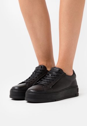 Černé kožené platformové tenisky nošené na bosých nohou, zobrazené z přední strany na bílém pozadí.