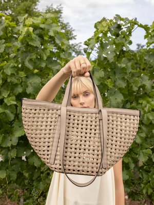 Mujer con cabello rubio sosteniendo un gran bolso tote tejido de color beige al aire libre frente a plantas verdes y frondosas.