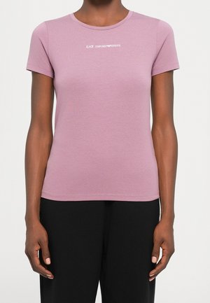 Personne portant un t-shirt rose ajusté à manches courtes avec le logo blanc EA7 Emporio Armani, associé à un pantalon noir, debout devant un fond uni.