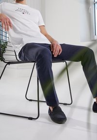 Muž v bílém tričku Tommy Hilfiger, námořnických kalhotách a černých botách sedí na černé drátěné židli v jasné místnosti s oknem.