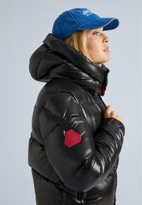 Donna in giacca a vento nera con colletto alto e patch rosso, indossa un cappellino da baseball blu e guarda verso destra contro uno sfondo semplice.