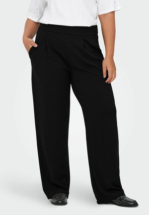 Schwarze weit geschnittene Hose mit flachem Bund, Falten und elastischem Bund. Kombiniert mit schwarzen Schuhen mit dekorativen Akzenten.