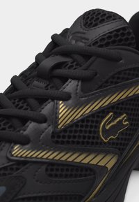 Μαύρο αθλητικό παπούτσι με πλέγμα ύφασμα, μαύρα κορδόνια και χρυσές ριγέ λεπτομέρειες, με χρυσό λογότυπο κροκόδειλου στην πλευρική επιφάνεια.