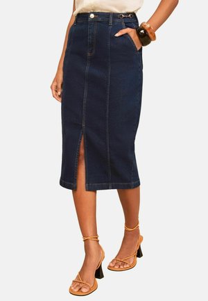 Kvinde iført en mørkeblå denim-midi nederdel med frontslids, hvid top, orange stropper på hælene og to store armbånd på håndleddet.