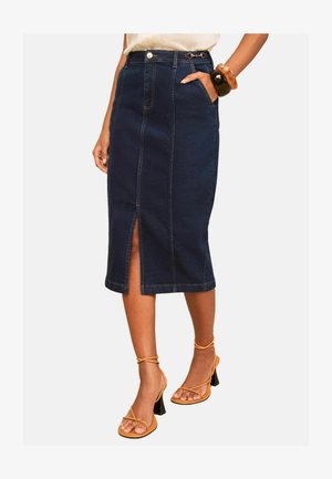Kvinde iført en mørkeblå denim-midi nederdel med frontslids, hvid top, orange stropper på hælene og to store armbånd på håndleddet.