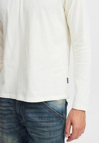 Langærmet cremefarvet bomulds skjorte med rundet kant, udstyret med et mærke på siden, parret med blå denim jeans.