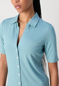 Polo bleu clair avec un bouton à l'avant, des manches courtes et un col. Texture lisse et design ajusté.