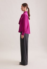 Blusa bordeaux con colletto annodato, maniche lunghe a sbuffo, abbinata a pantaloni grigi a vita alta e scarpe nere a punta.