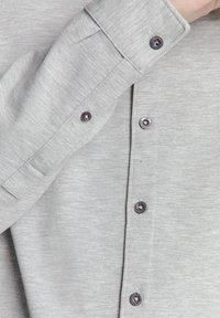 Grijze lange mouwen shirt met een zachte textuur, voorzien van een rij zwarte knopen en gestructureerde manchetten. De stof is subtiel getextureerd.