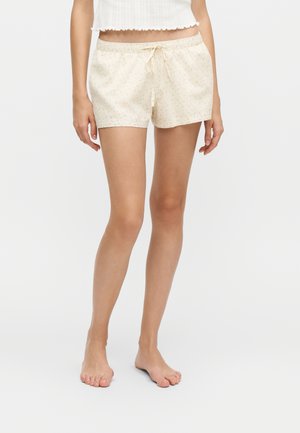 Frau trägt cremefarbene Shorts mit kleinem gepunktetem Muster und Kordelzug, steht barfuß vor weißem Hintergrund.