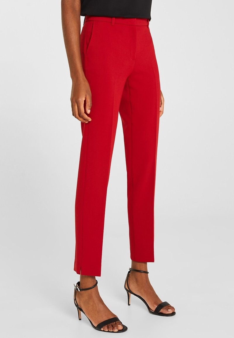 Femme portant un pantalon rouge sur mesure longueur cheville avec fentes latérales, associé à des sandales noires à talons hauts et à brides.