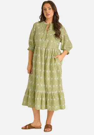 Frau in einem grünen, gemusterten Midikleid mit Puffärmeln und braunen Slip-On-Sandalen, stehend mit einer Hand in der Tasche.