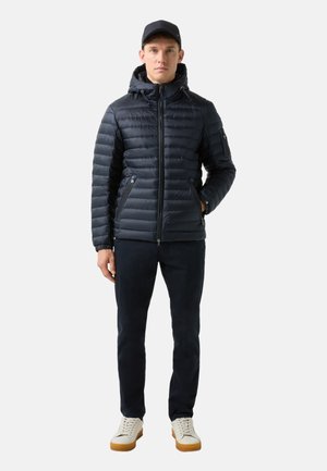 Marineblaue Daunenjacke mit Kapuze und Reißverschluss, ausgestattet mit horizontalen Steppnähten und seitlichen Taschen. Kombiniert mit dunklen Hosen und hellen Sneakern.