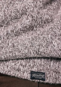 Detail šedé a bílé pletené látky s černou štítkem s nápisem "Heritage Best Jeans Quality Since 79" na hnědém oblečení pod ní.