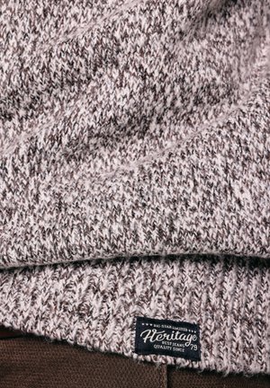 Tuvplāns pelēkas un baltas adītas auduma ar melnu etiķeti, uz kuras rakstīts "Heritage Best Jeans Quality Since 79", uz brūna apģērba zem tā.