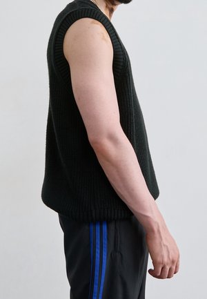 Vue de profil d'une personne portant un gilet tricoté noir sans manches et un pantalon noir à rayures bleues sur un fond uni.