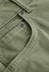 Un tissu vert olive avec une texture lisse présente deux boutons en métal noir et des coutures visibles le long des bords de la poche et de la patte de boutonnage.