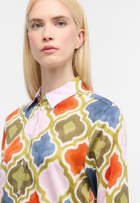 Farverig skjorte med et geometrisk mønster i orange, pink, blå og grøn. Har knaplukning foran og klassisk krave. Glat tekstur.
