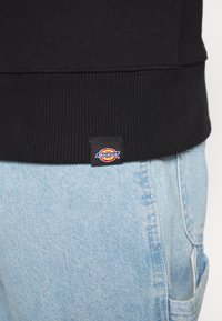 Felpa nera con orlo e polsini a coste, con un piccolo etichetta che mostra il logo "Dickies". Abbinata a jeans blu chiaro.