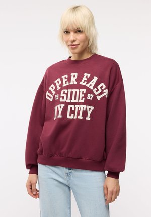Osoba o blond włosach, ubrana w bordową bluzę z napisem "Upper East Side 1997 NY City" i jasnoniebieskie dżinsy na białym tle.