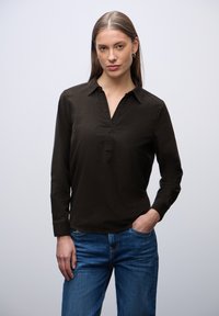 Camisa de color marrón oscuro con cuello y una tapeta de botones parcial, combinada con unos vaqueros de denim azul. Textura suave, mangas largas, corte relajado.