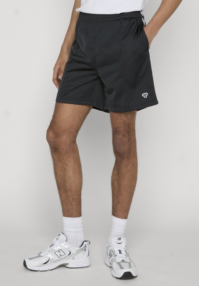 Homme portant un short de sport noir, des chaussettes blanches montant jusqu'au mollet et des baskets blanches New Balance, debout avec une main dans la poche.