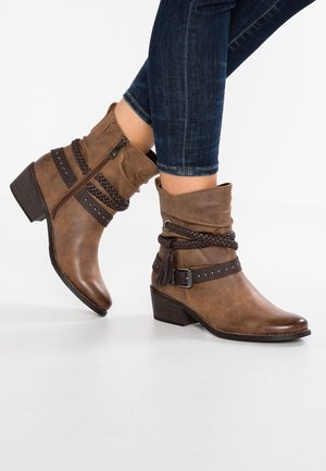Brown leather ankle boots with braided straps, buckles, side zipper, worn with dark blue skinny jeans, against a white background.