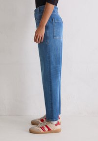 Personne portant un jean droit bleu et des baskets beiges avec des rayures rouges, se tenant contre un mur uni de couleur claire.