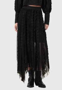 Midi φούστα από μαύρη δαντέλα με κυματιστά κάθετα πάνελ, διαφανές ύφασμα, σετάρισμένη με μαύρα μποτάκια αστραγάλου και μακρυμάνικη μπλούζα, τα χέρια χαλαρά στο πλάι.