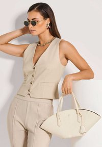 Beige maßgeschneiderte Weste mit tiefem V-Ausschnitt und Knopfverschluss, kombiniert mit passenden Hosen; hält eine weiße, asymmetrische Handtasche.