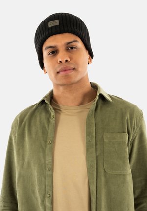 Un bonnet côtelé noir, une chemise en velours côtelé verte avec une poche en patch, et un t-shirt beige. Les textures et couleurs des tissus contrastent pour un intérêt visuel.
