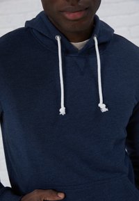 Marineblauwe hoodie met een trekkoordkap, V-halsdetail en voorzak; gemaakt van zachte stof met een gladde textuur en witte accenten.