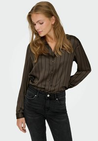 Blusa de seda verde oscuro con rayas negras verticales, mangas largas y cierre de botones en la parte delantera, combinada con jeans negros. Textura y ajuste elegantes.