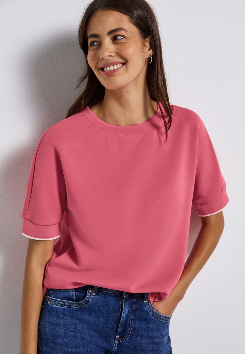 Haut rose à manches courtes avec un col rond, présentant des bordures blanches sur les manches. Le tissu semble lisse, porté avec un jean en denim bleu.