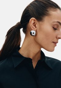 Orecchini sferici color argento metallizzato, finitura lucida, indossati sull'orecchio con una camicia nera con bottoni, capelli raccolti. Sfondo neutro.