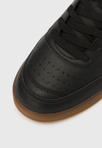 Crni kožni vrh tenisice s perforacijama, crnim vezicama i smeđim gumiranim đonom na bijeloj pozadini.