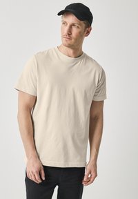 Béžové bavlněné tričko s krátkým rukávem, kulatým výstřihem a volným střihem, spárováno s černými džíny a černou kšiltovkou.