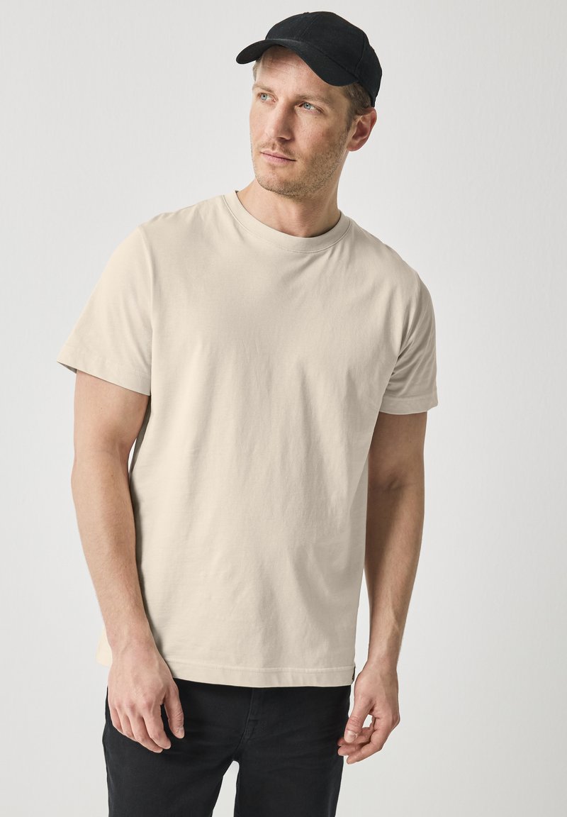 Béžové bavlněné tričko s krátkým rukávem, kulatým výstřihem a volným střihem, spárováno s černými džíny a černou kšiltovkou.