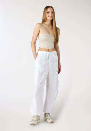 Giovane donna con capelli lunghi e lisci che indossa un top beige senza spalline, pantaloni bianchi ampi e sneakers beige, in piedi con le mani nelle tasche.