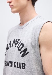 Νεαρό άτομο που φοράει γκρι αμάνικο μπλουζάκι με την επιγραφή "CHAMPION RUNNING CLUB" σε μαύρο κείμενο στο στήθος.
