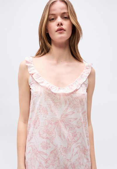 Νεαρή γυναίκα φορώντας ένα αμάνικο ανοιχτό ροζ φόρεμα με λουλούδια και στρογγυλεμένο λαιμό με βολάν, στέκεται μπροστά σε ένα απλό φωτεινό φόντο.