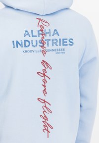 Világoskék kapucnis pulóver, puha anyagból készült, piros és kék színű "ALPHA INDUSTRIES" felirattal és szlogennel a hátsó részen.